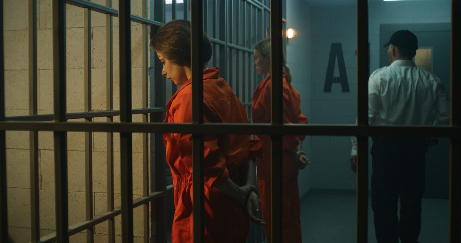 Two female prisoners, inmates in orange uniforms stand facing the metal bars in front of prison cells as an officer walks by. prison inspection - federal prrison for women - FCI Tallahassee