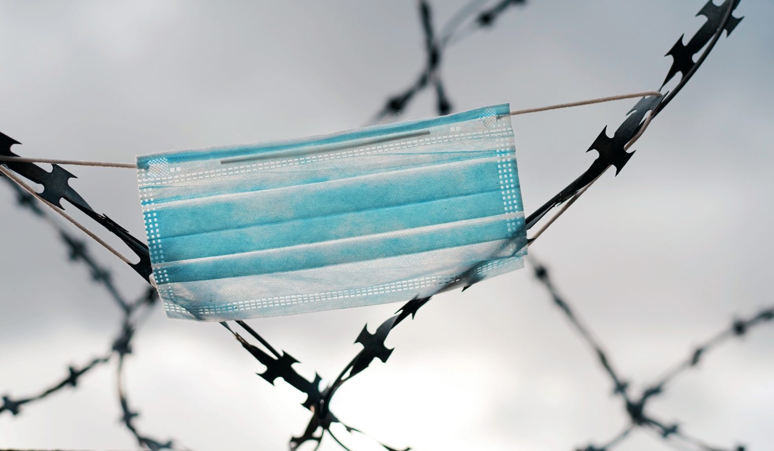 A medical mask hanging on barbed wire. News - Impact of 2021 Tuberculosis in Prison Outbreak Persists, Prompts Health Department Advisory